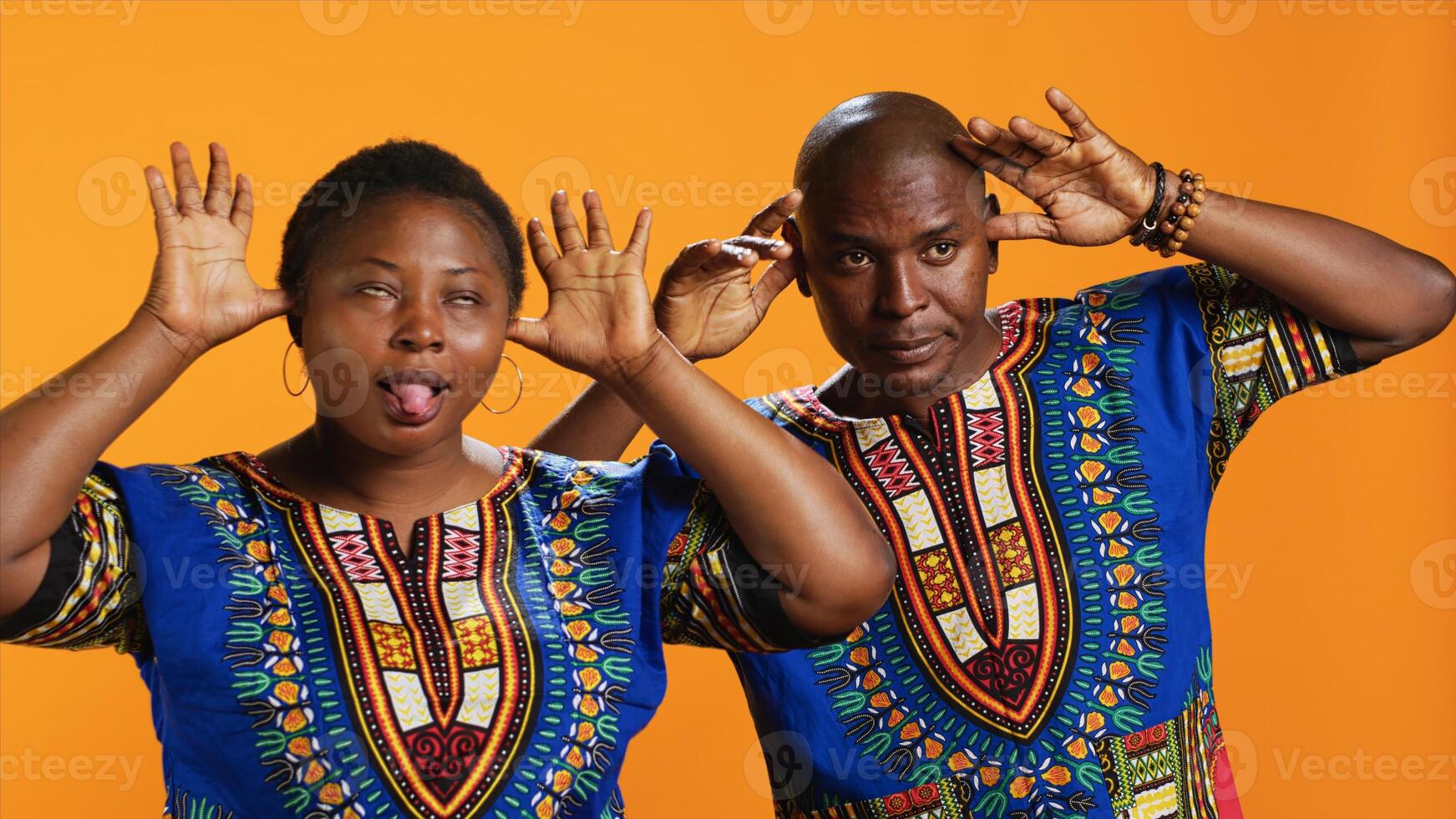 Silly ethnic couple fooling around on camera with comic face, having fun and sticking tongue out in studio. Cheerful people doing funky gesture and enjoying time on set, orange background. photo