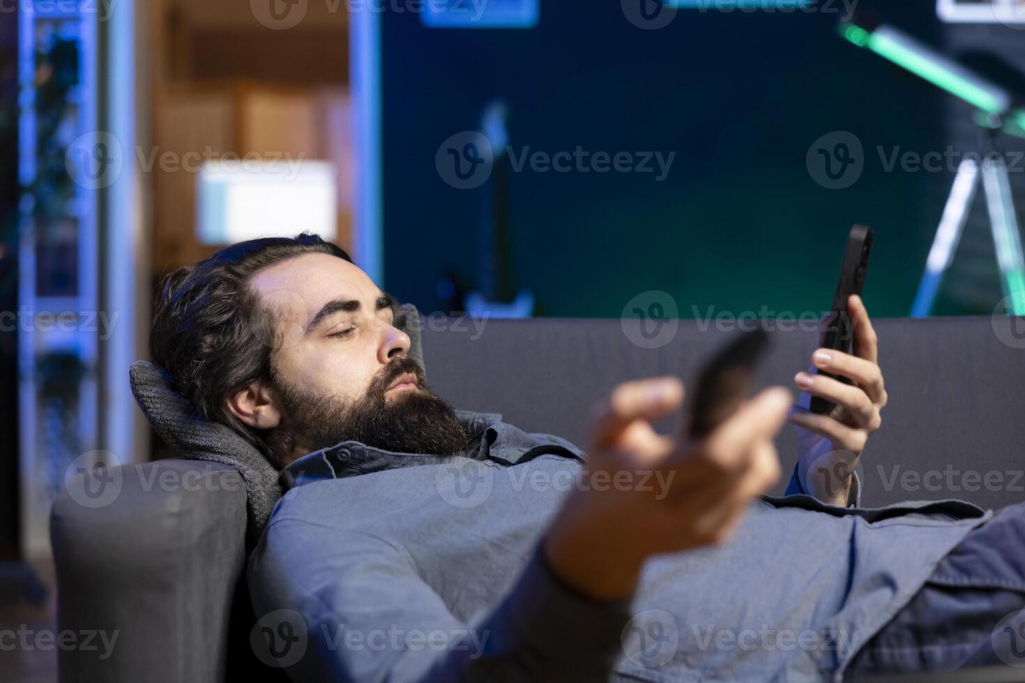 Homeowner bored at home laying down, using TV to watch movies on streaming services and chatting with friends. Apathetic man lounged on couch, mindlessly consuming entertainment and using mobile phone photo