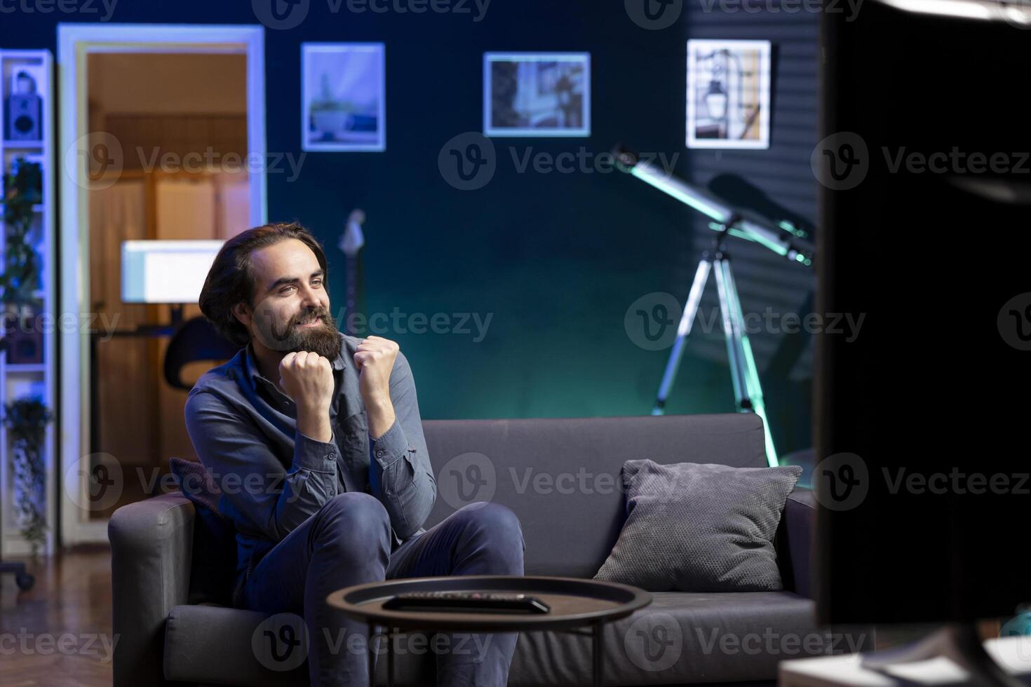 Happy man watching live sports event broadcasted on TV, rooting for favorite team. Joyous supporter sitting on couch at home enjoying football competition, celebrating win photo