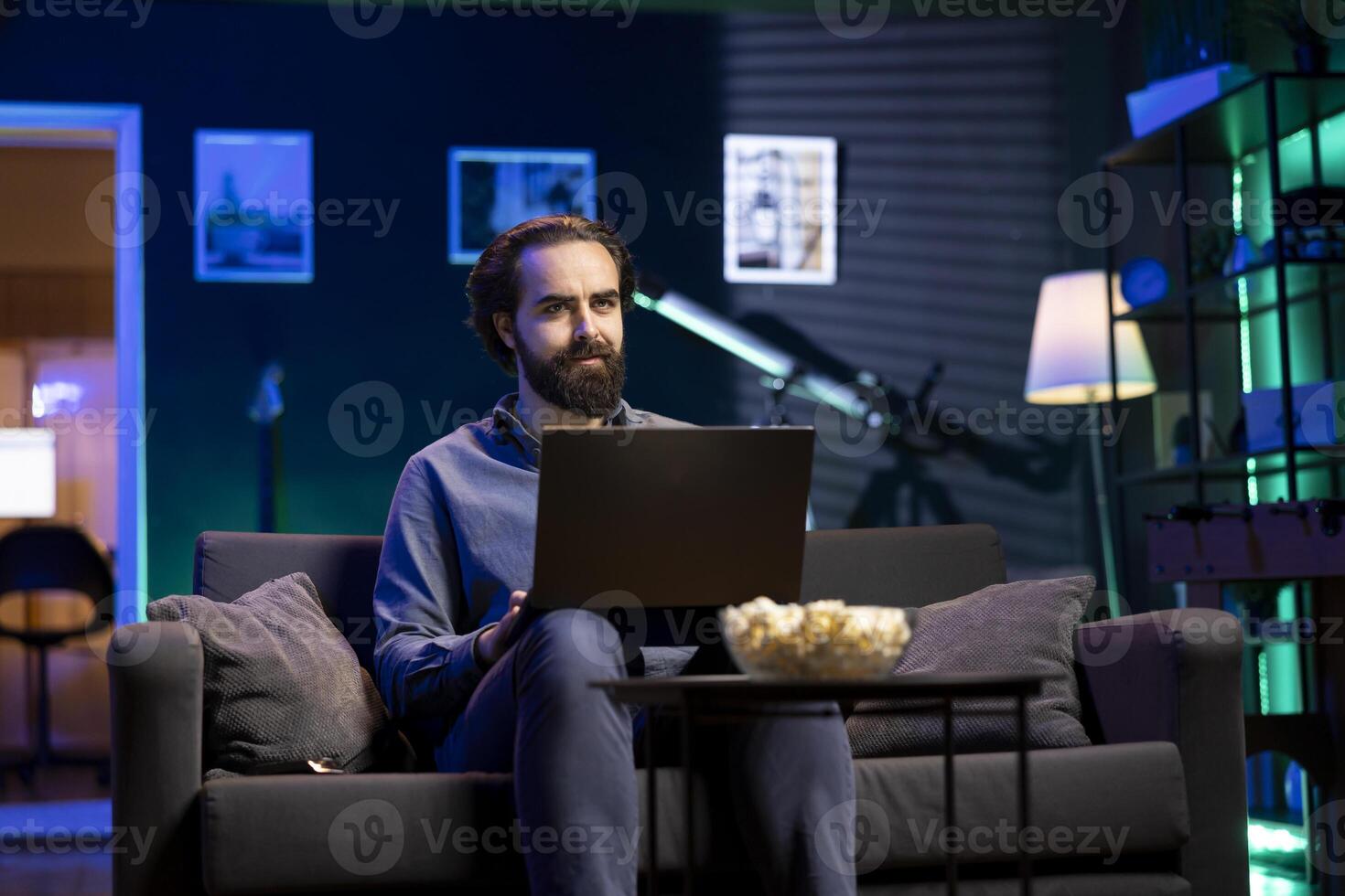Man enjoying bowl of popcorn while watching cable programs on TV and sending emails on laptop. Spectator on couch in living room eats snacks during movie and works on notebook photo