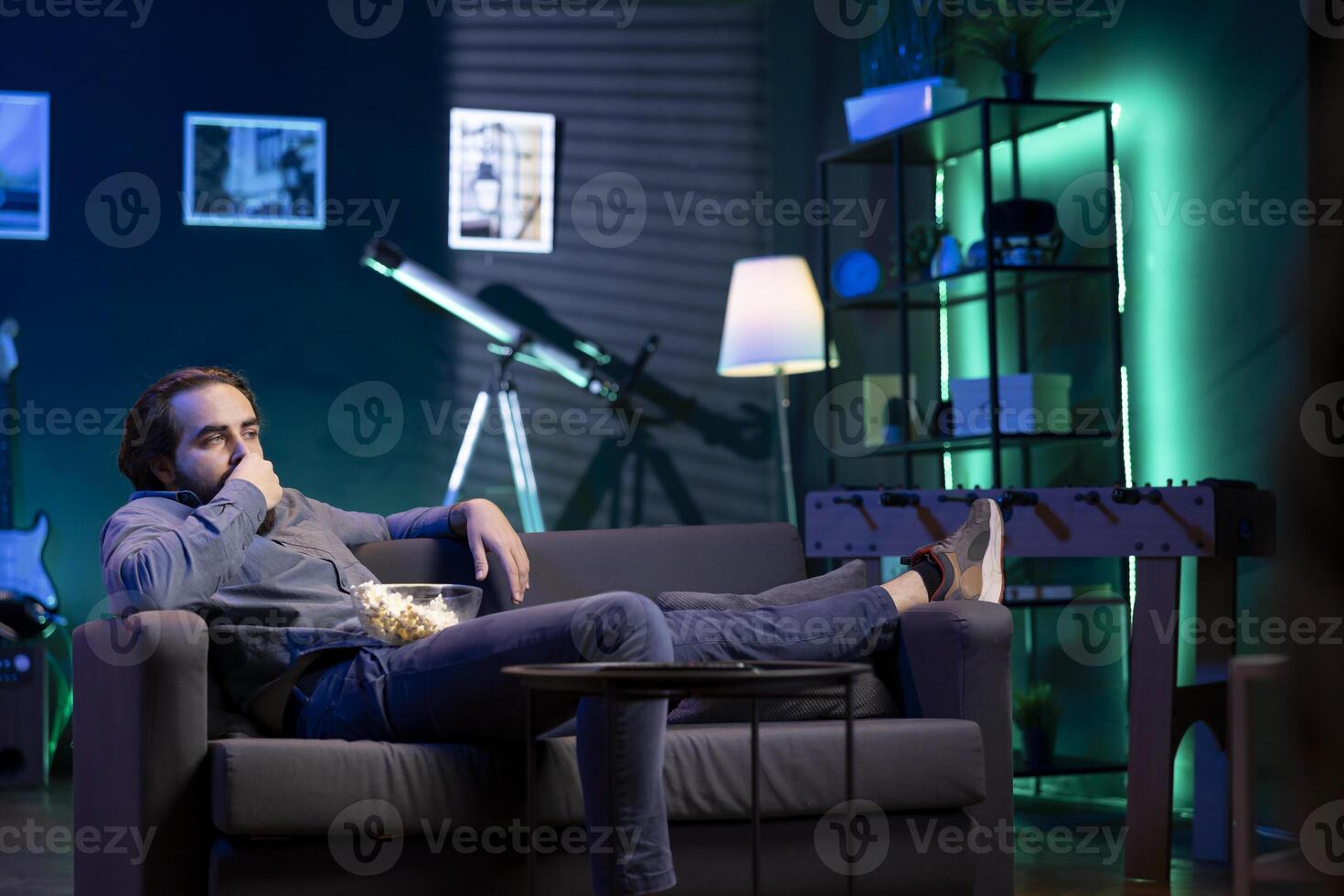 Homeowner bored at home laying down, using TV to watch movies on subscription based streaming services. Apathetic man lounged on couch, mindlessly consuming entertainment, eating popcorn photo