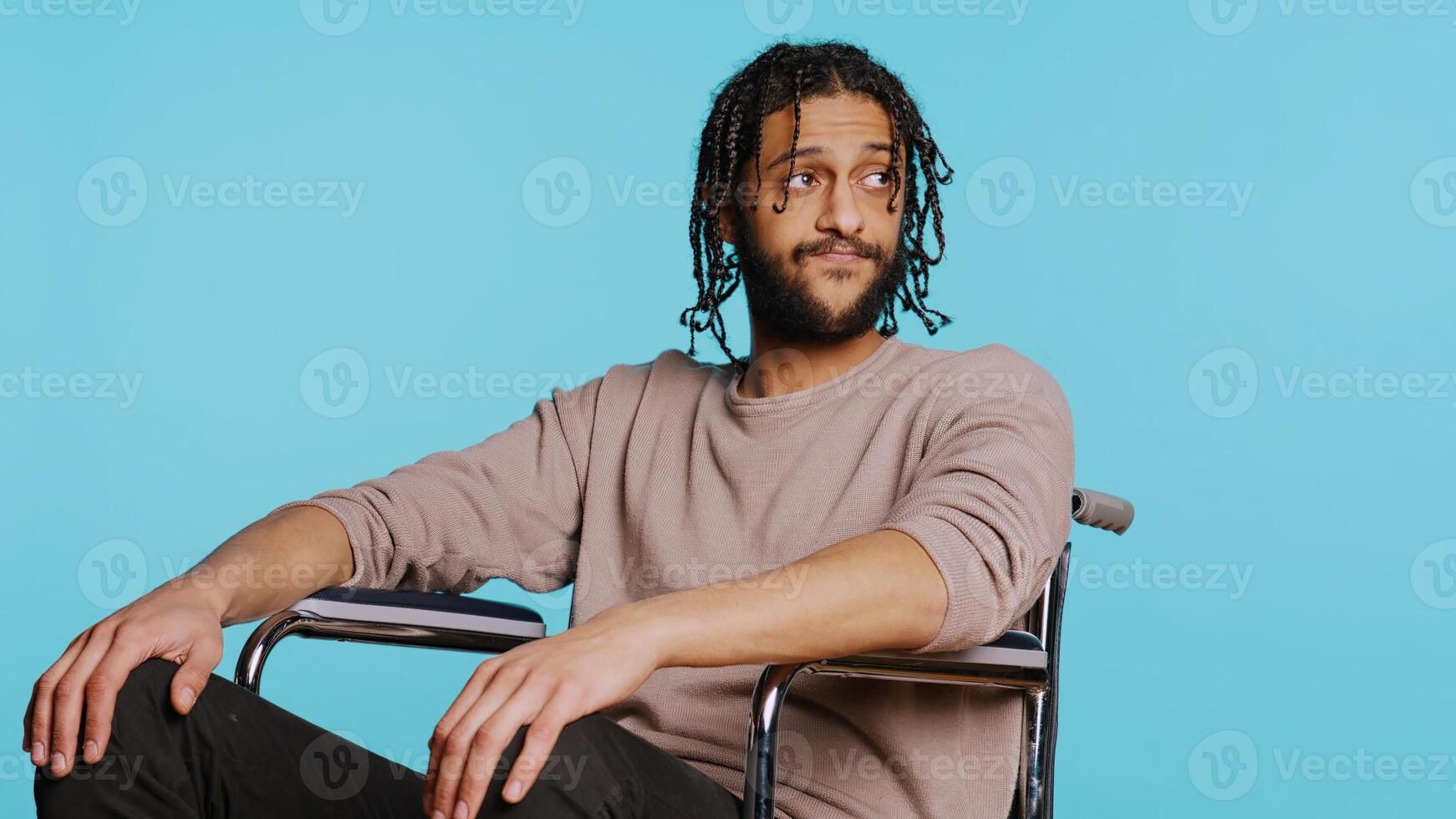 Bored Middle Eastern man with disability in wheelchair impatiently tapping fingers on knee, waiting. Annoyed person with paraplegia looking around, isolated over studio background, camera B photo