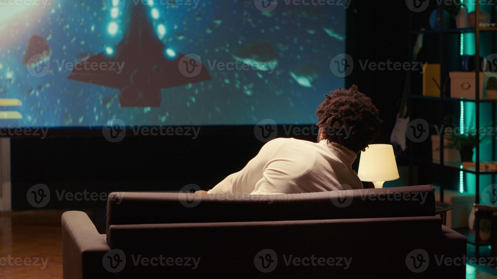 African american man plays science fiction singleplayer game with overlay HUD showing health bars and minimap on huge TV screen. Gamer enjoying high quality ray tracing graphics in console videogame photo