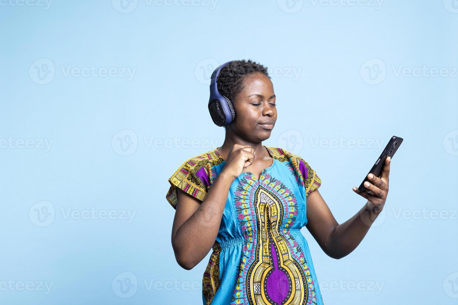 Female model playing music from a playing in the studio, having fun with groovy music on audio headset over blue background. Smiling confident woman listening to tunes on headset. photo