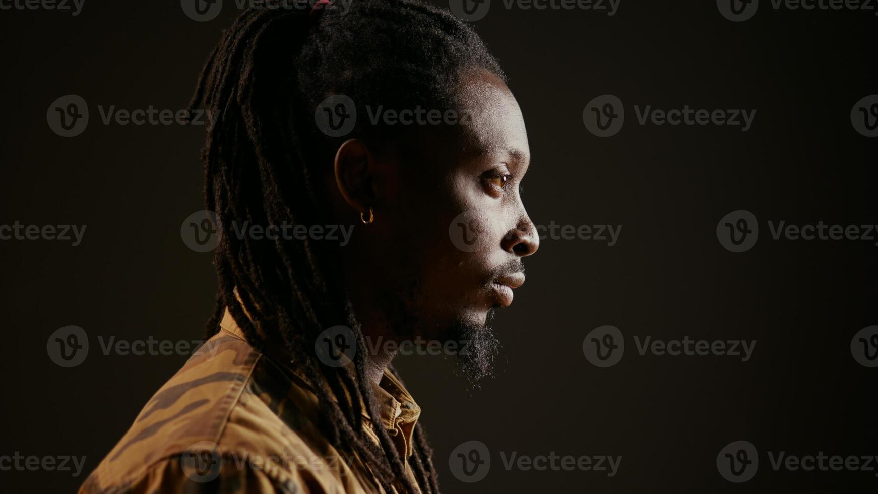 African american guy looking at camera with confidence, feeling happy and relaxed with dreads and cool clothing items. Model posing, standing over black background smiling. Handheld shot. photo