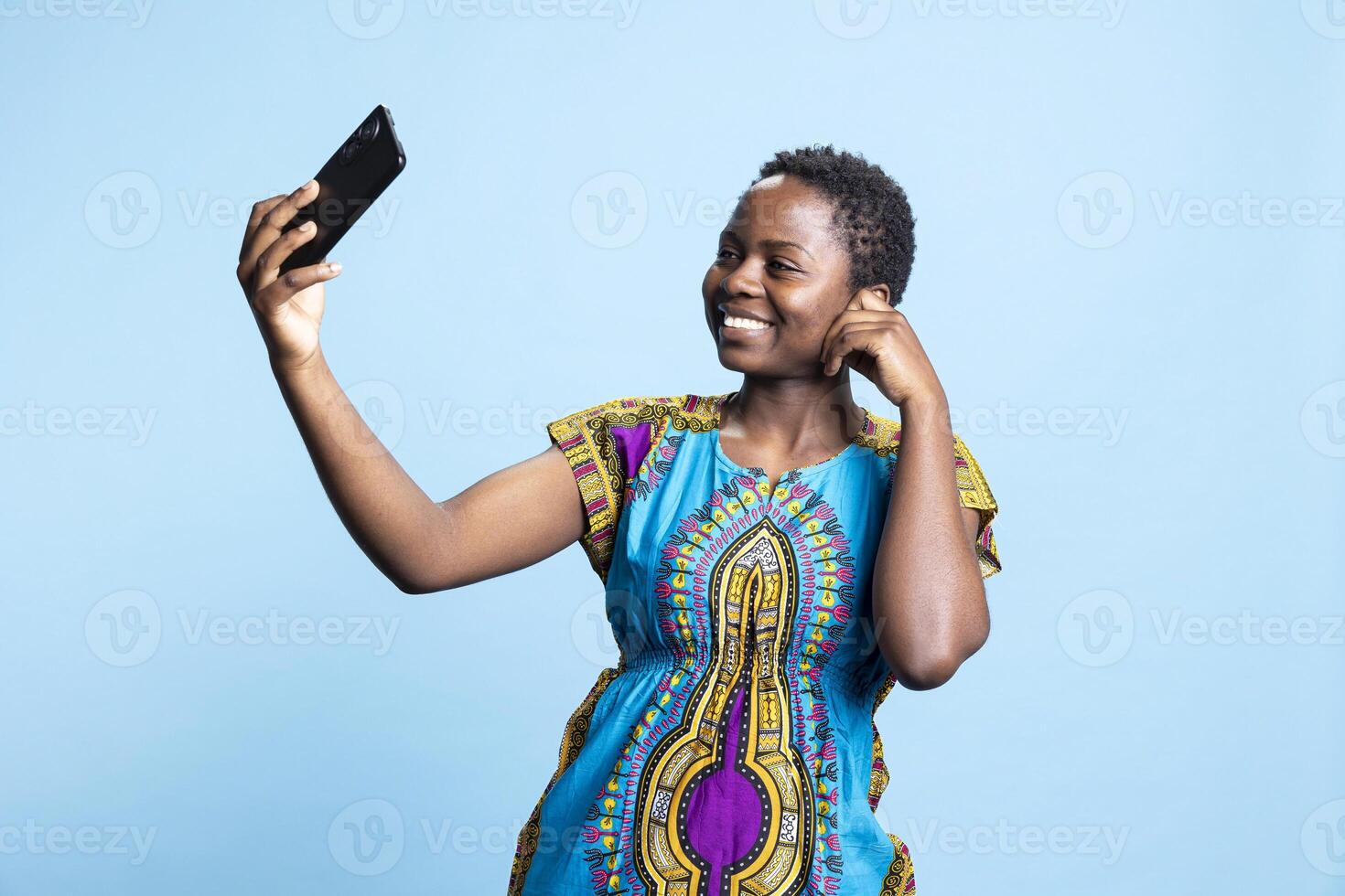 African american woman smiling at smartphone camera in studio, taking pictures and posting on social media apps. Female model photographing herself over blue background, ethnic style. photo