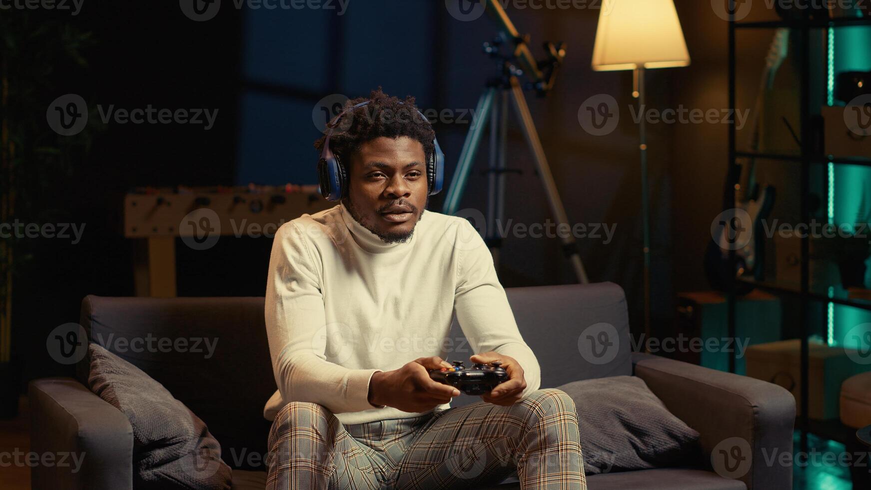 Man in rgb lit apartment playing games on couch, enjoying day off from work. Gamer battling enemies in online multiplayer shooter on gaming console, talking with teammates through headphones photo