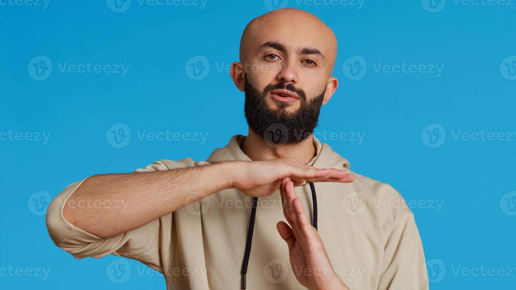 Middle eastern person doing timeout gesture on camera, asking for a break to stop working over blue background. Muslim adult presents symbol to pause or stop something. Camera 2. Handheld shot. photo