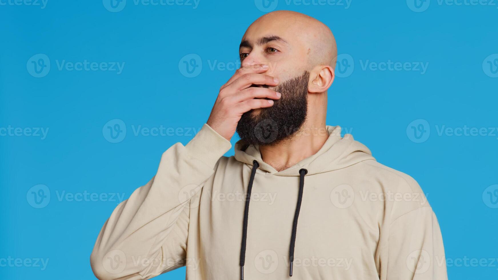 Middle eastern man feeling exhausted and yawning in studio, covering his mouth in front of the camera. Arab person being sleepy in the morning, awake from the early hours. Camera 2. photo