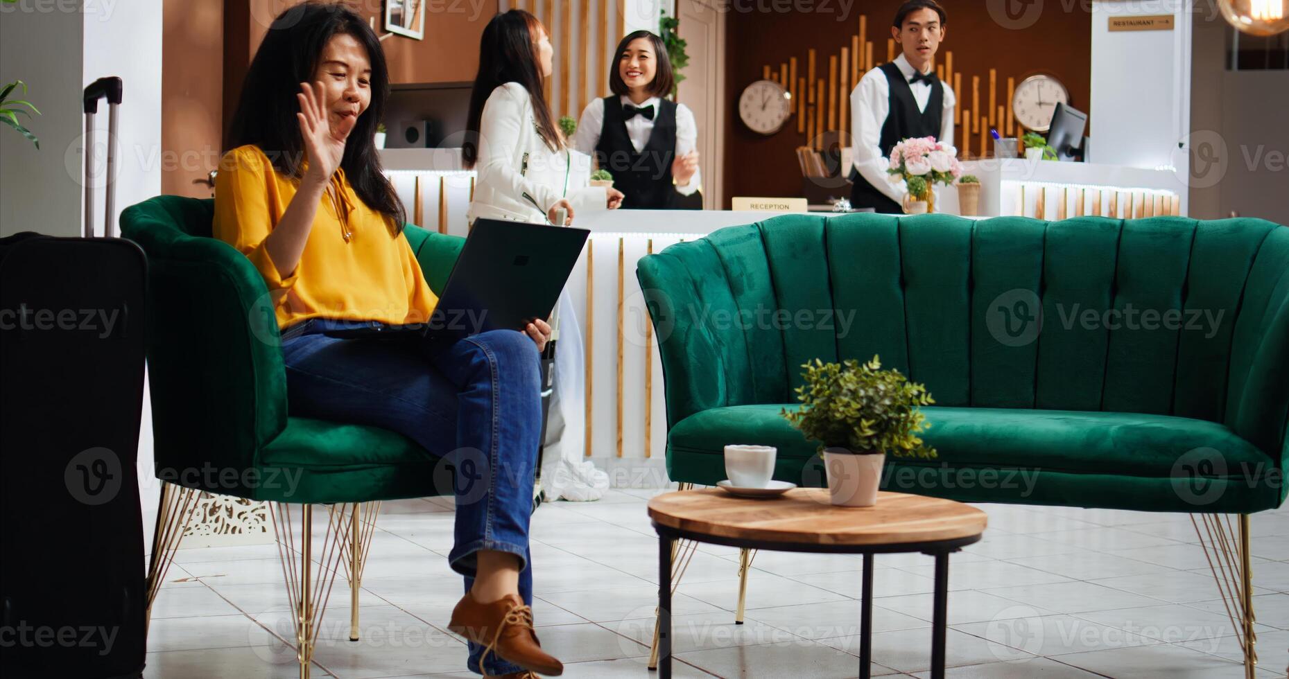 Asian tourist chats on online laptop videocall at hotel, before registering she is talking to family after arriving from the airport. Woman using remote videoconference connection in lounge area. photo