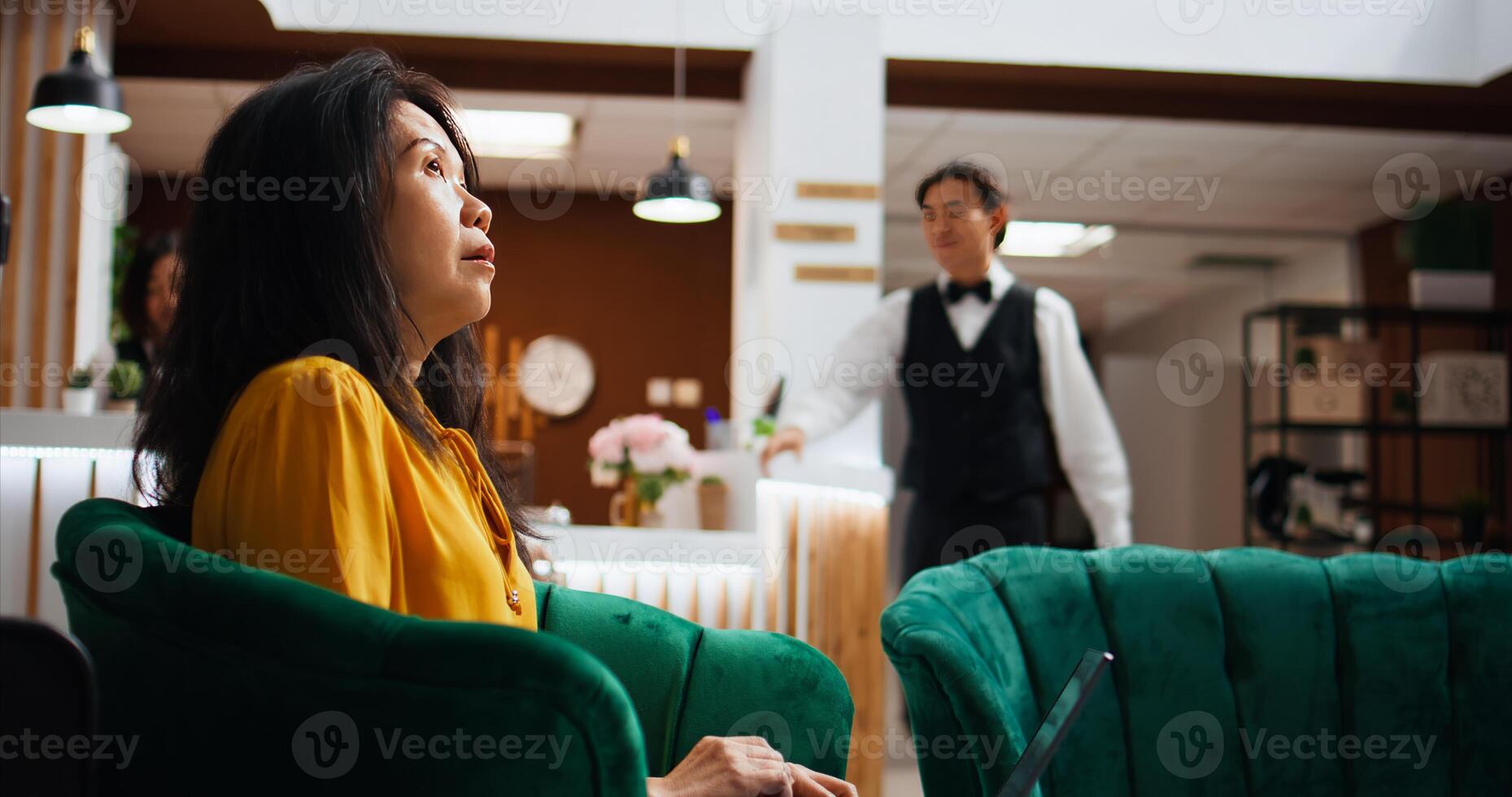 Hotel client solving issues on laptop in lounge area, waiting to register and see accommodation. Asian tourist passing time on pc, waiting to be called at front desk reception. Handheld shot. photo