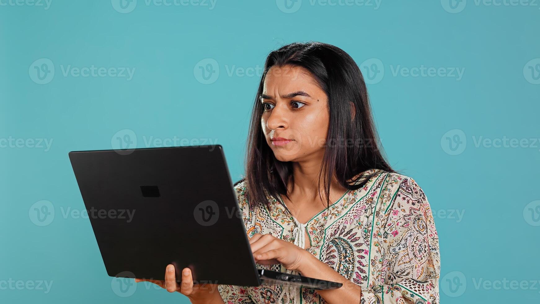 Indian woman typing on laptop keyboard and cursing, feeling angry at work. Person writing emails on notebook, complaining loudly, isolated over studio background, camera B photo