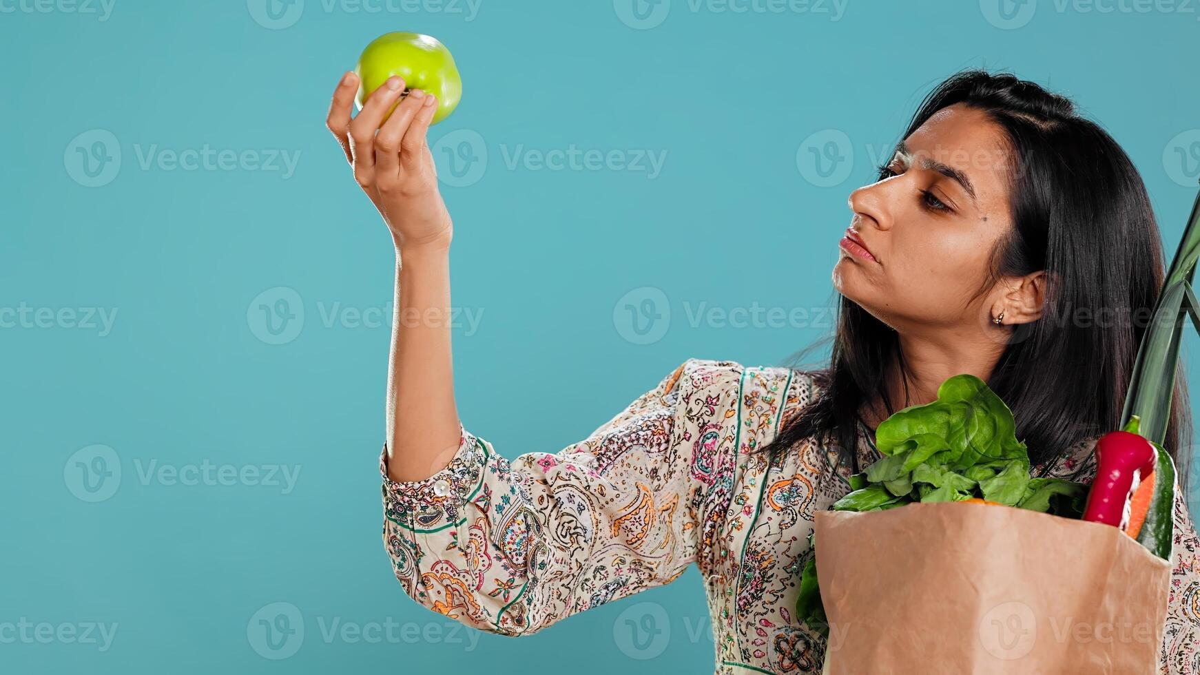 Indian woman with reusable paper bag in hands looking at apple, living healthy lifestyle. Ecology lover holding shopping bag with natural produce, studio background, camera B photo