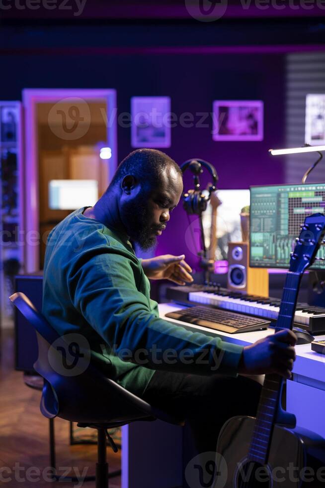 African american artist playing guitar and electronic piano synthesizer in home studio, recording acoustics with mixing console to produce cool tunes. Sound technician editing audio recorded files. photo