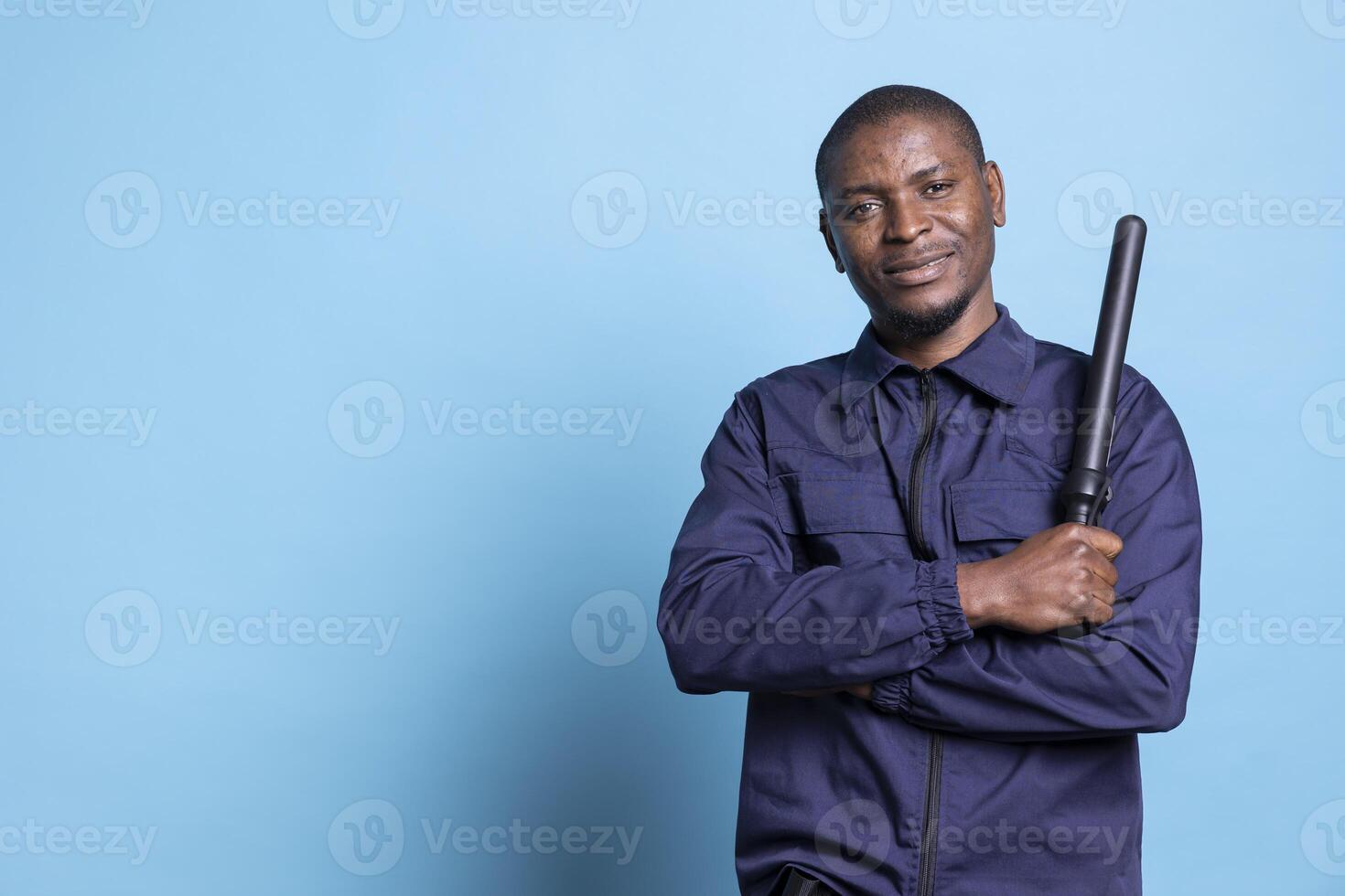 African american security officer feels proud with his duty in studio, posing with confidence holding his baton dressed in professional uniform. Skilled bodyguard standing against blue background. photo