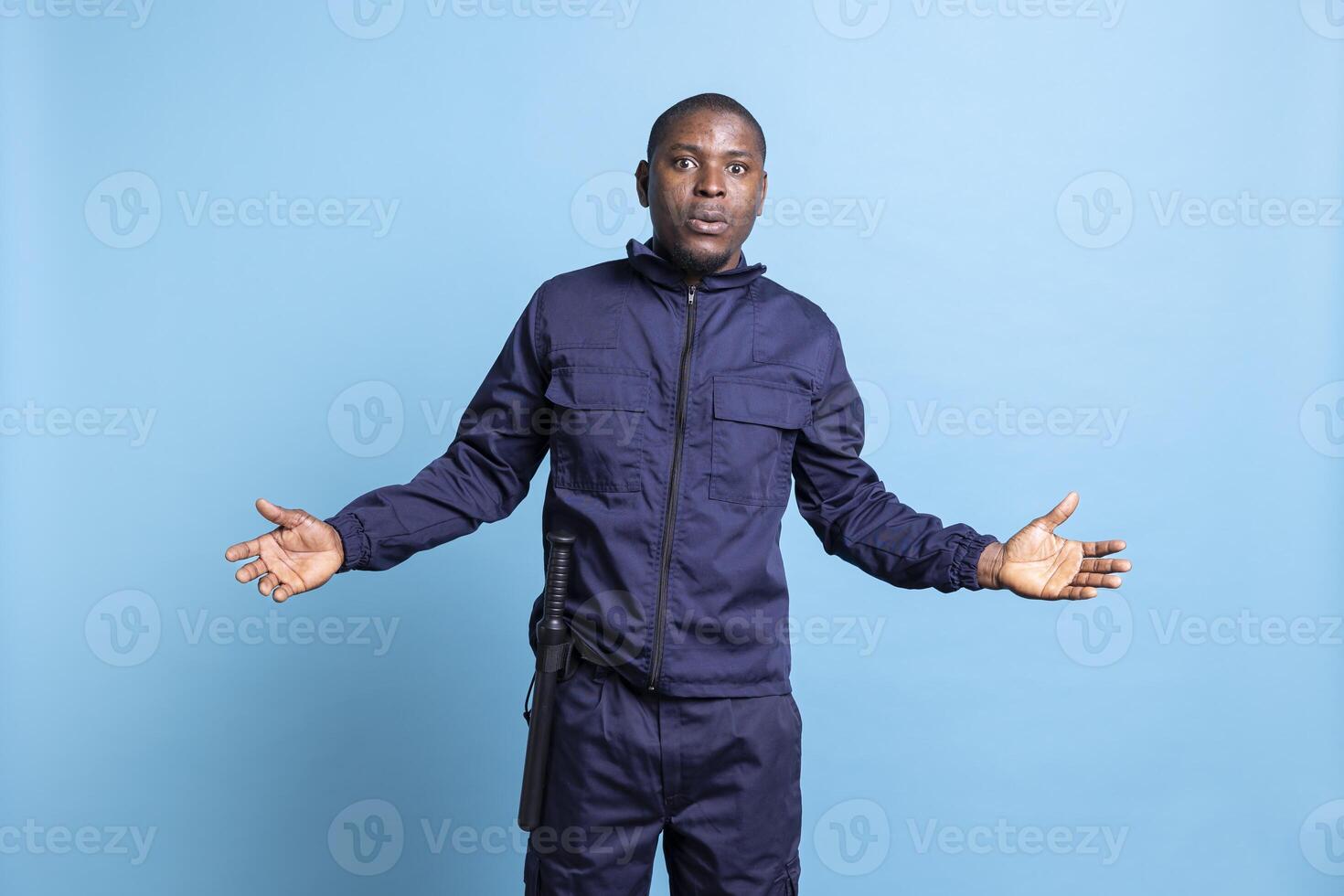 Security guard in uniform is acting surprised in front of the camera, being in shock after finding out something. African american protection agent with baton feels amazed against blue background. photo