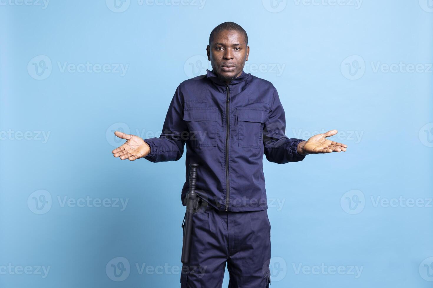 Clueless unsure security officer shrugging against blue background, raising his shoulders and showing i dont know sign. Bodyguard acting uncertain and doubtful, confused personnel. photo