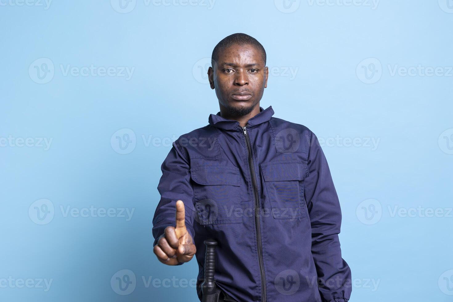 African american security guard poses with a finger up in studio, presenting a restriction sign against blue background. Person working as a patrol officer showing a stop symbol in studio. photo