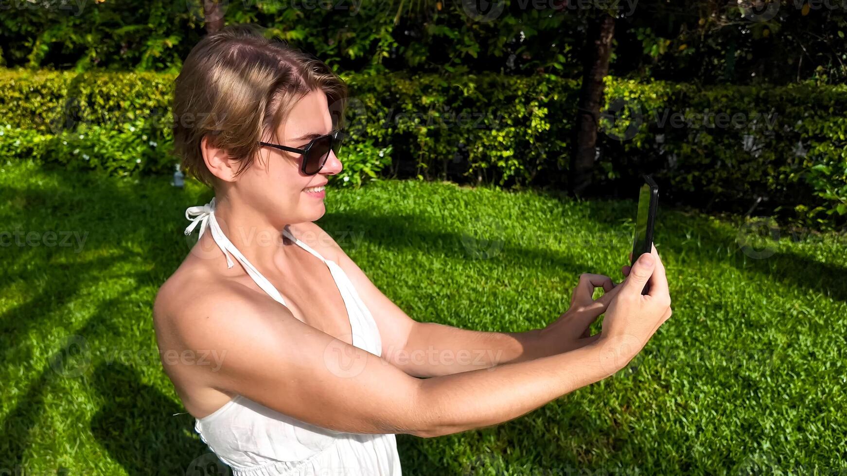 Adult Caucasian woman in sunglasses using smartphone in sunny park, symbolizing digital connectivity and leisure during summer vacations photo