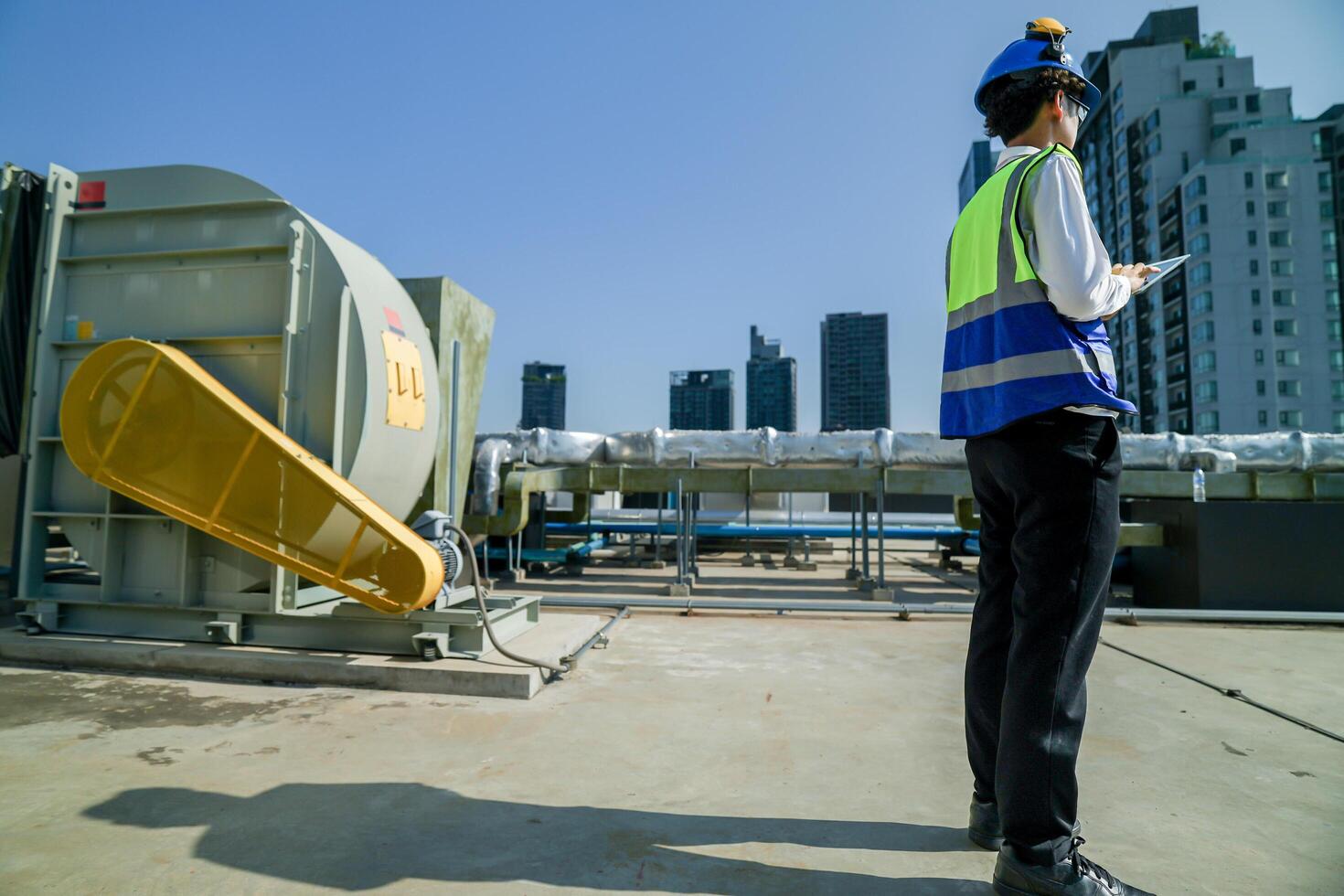 Skilled engineer operating tablet to manage HVAC machinery on top of building, urban architecture in distance. photo