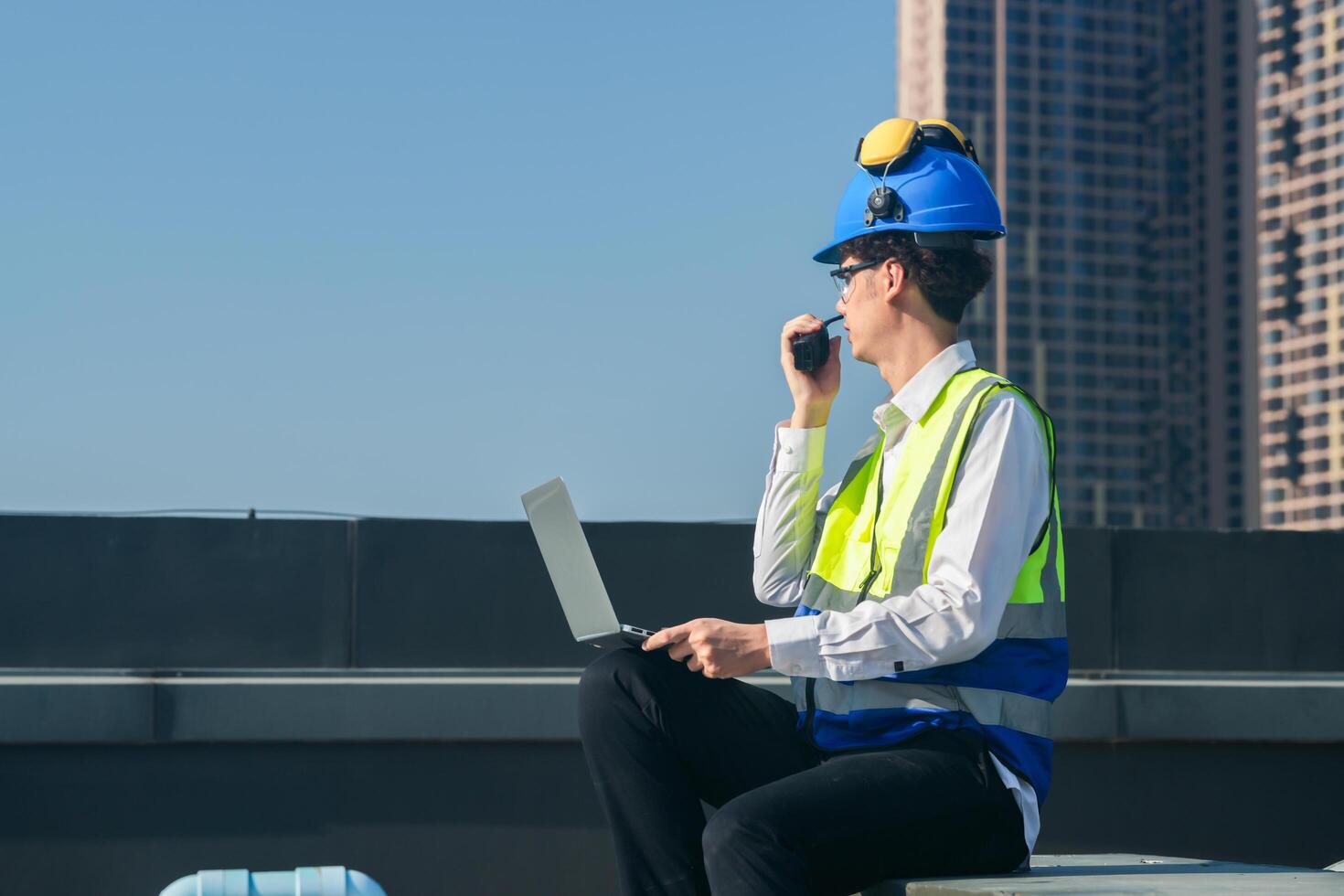 Focused engineer in hard hat and safety vest using walkie-talkie and laptop on construction site with urban backdrop. photo