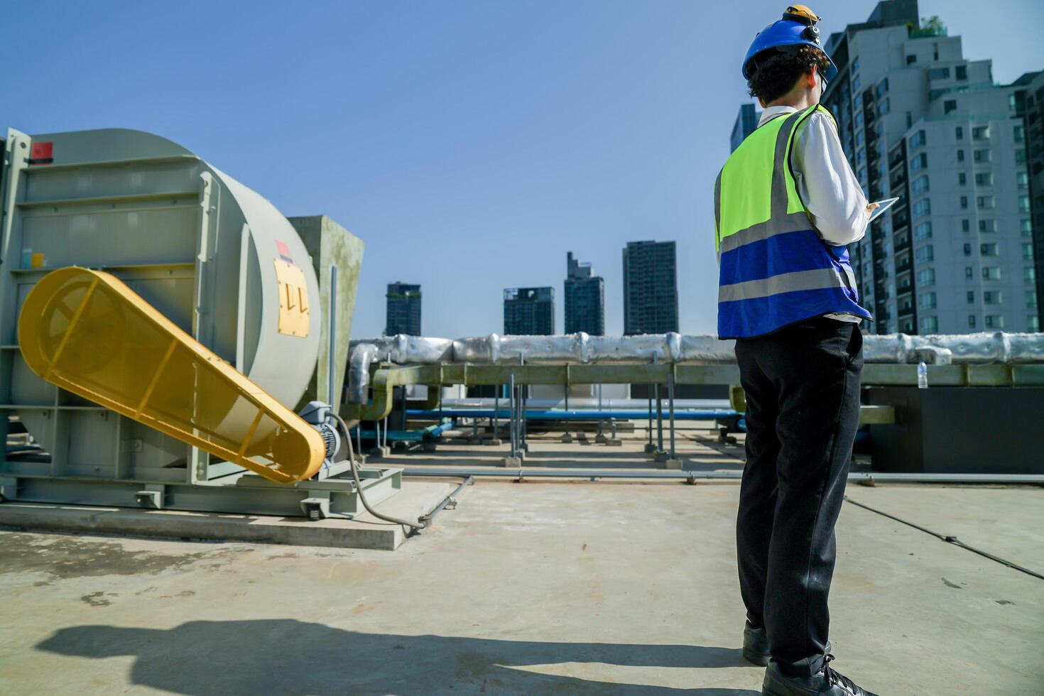 Skilled engineer operating tablet to manage HVAC machinery on top of building, urban architecture in distance. photo