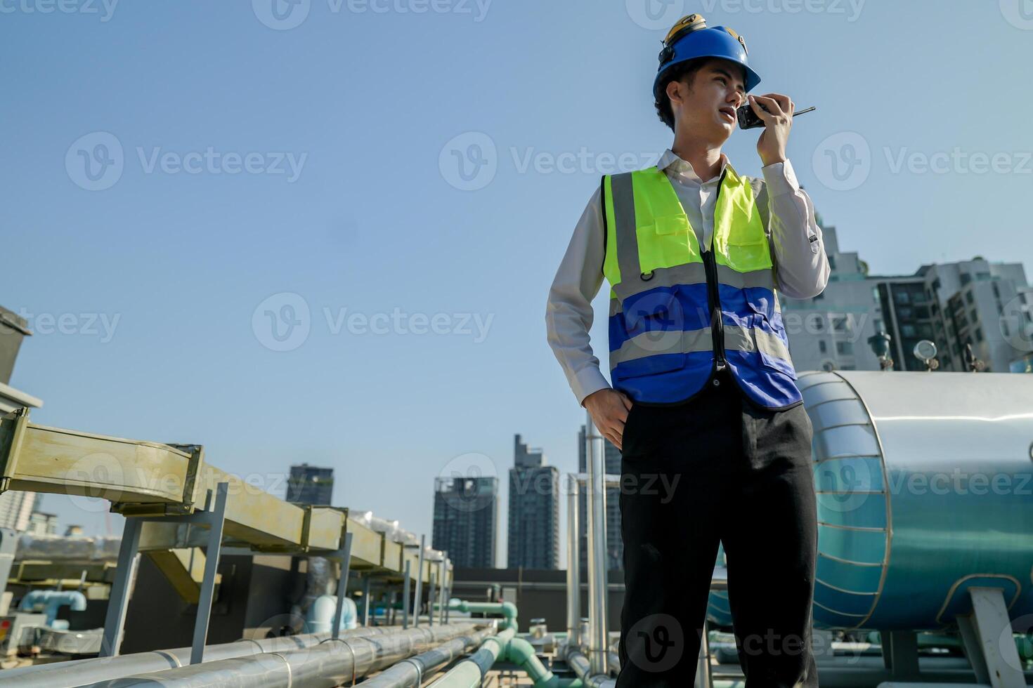 A young engineer checks equipment on a rooftop amidst urban skyline, clipboard in hand, ensuring system functionality.Engineer in safety vest and helmet conducting maintenance on rooftop machinery. photo