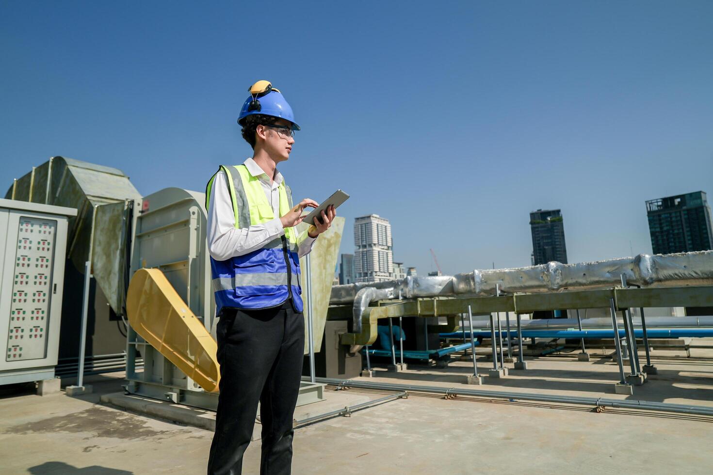 Engineer with tablet on rooftop assessing HVAC system against city skyline, safety equipment and hard hat on. photo
