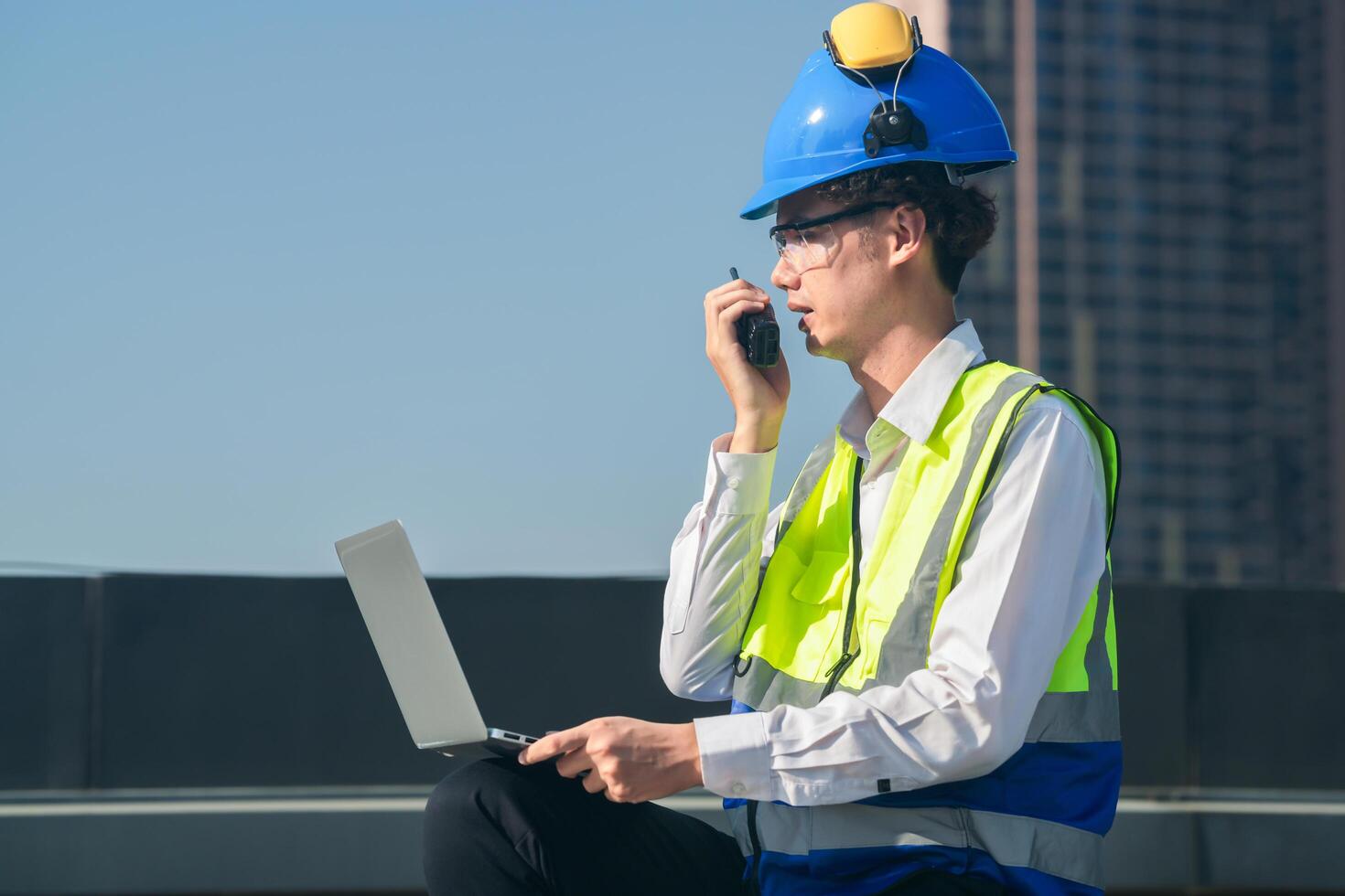 Focused engineer in hard hat and safety vest using walkie-talkie and laptop on construction site with urban backdrop. photo