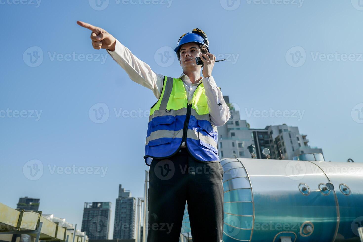 A young engineer checks equipment on a rooftop amidst urban skyline, clipboard in hand, ensuring system functionality.Engineer in safety vest and helmet conducting maintenance on rooftop machinery. photo