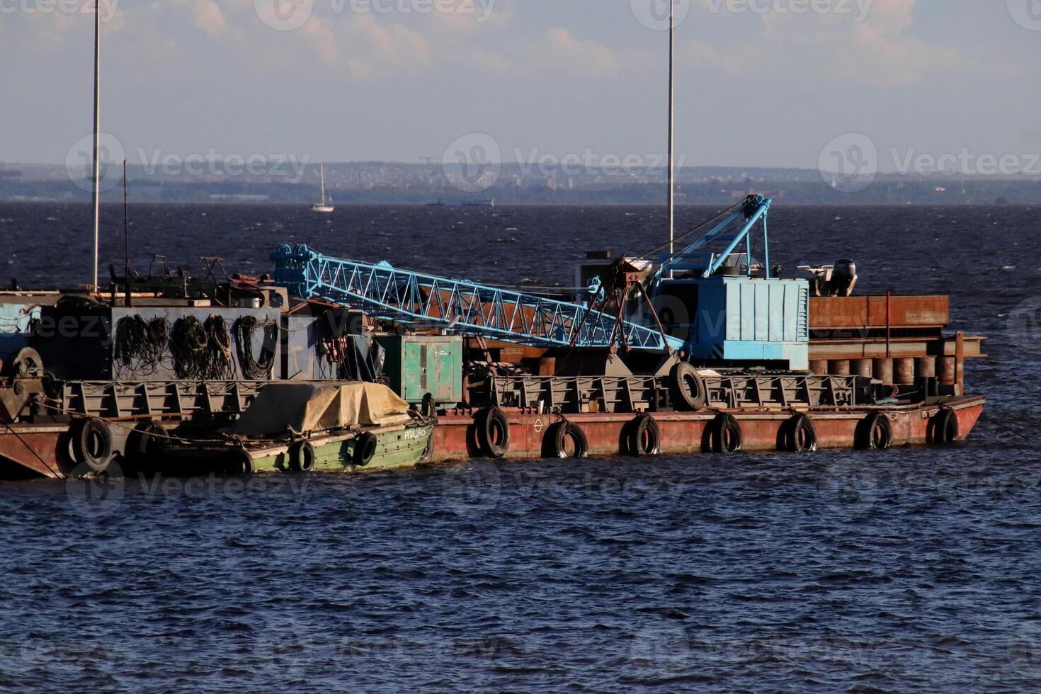 A pontoon with a crane and rusty superstructures floats near the pier on the shore. photo