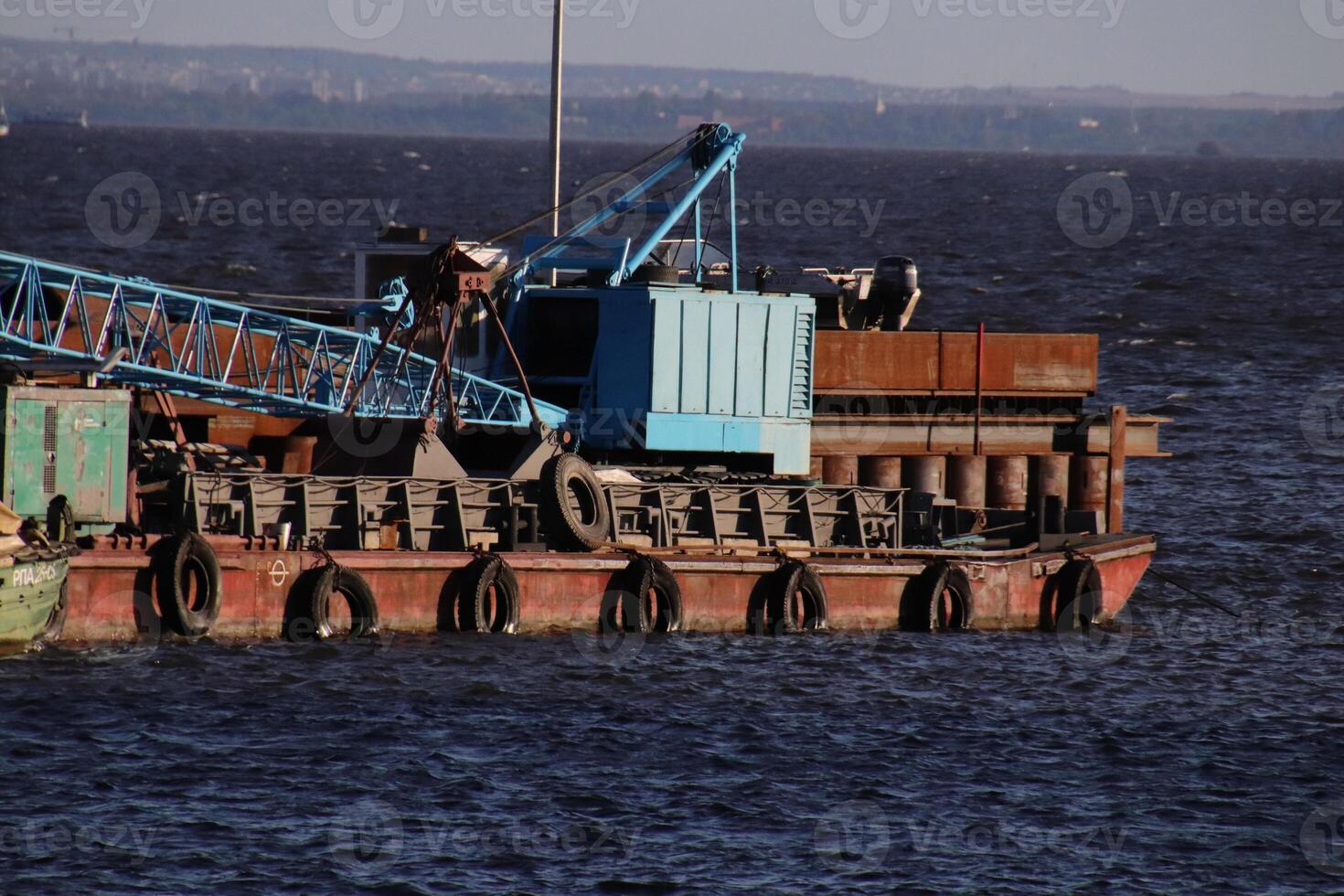 A pontoon with a crane and rusty superstructures floats near the pier on the shore. photo