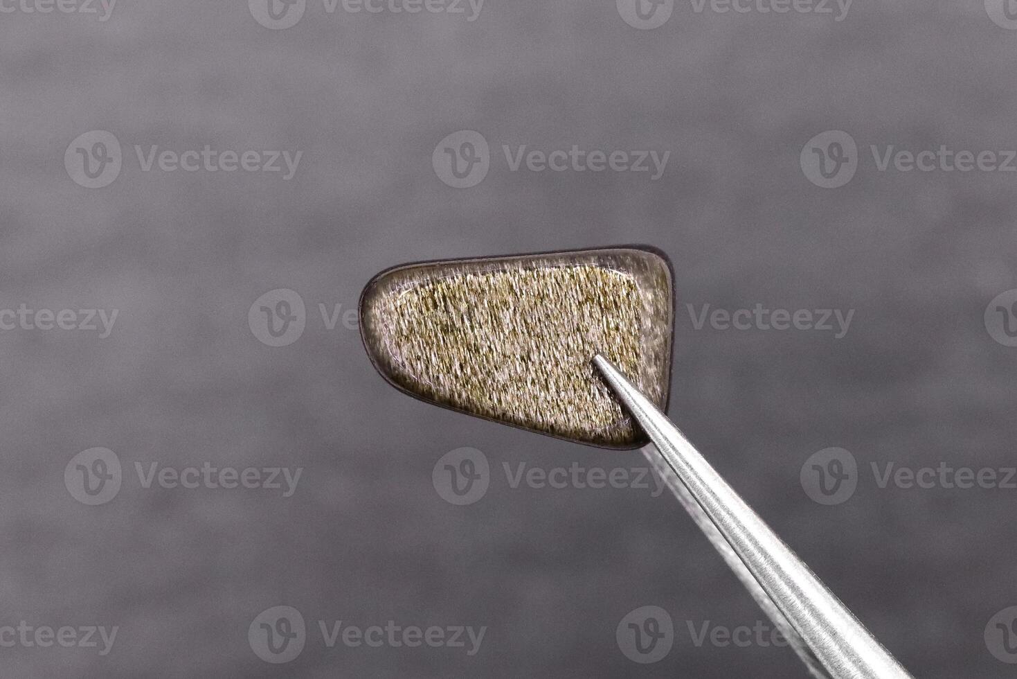 Gold obsidian jewel stone texture in tweezers on gray background. Macro closeup. photo