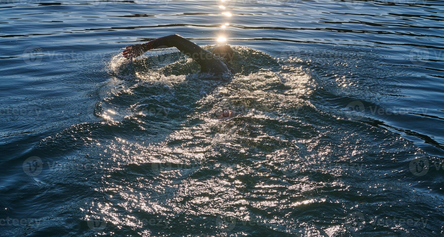 Triathlon athlete swimming on lake in sunrise wearing wetsuit photo