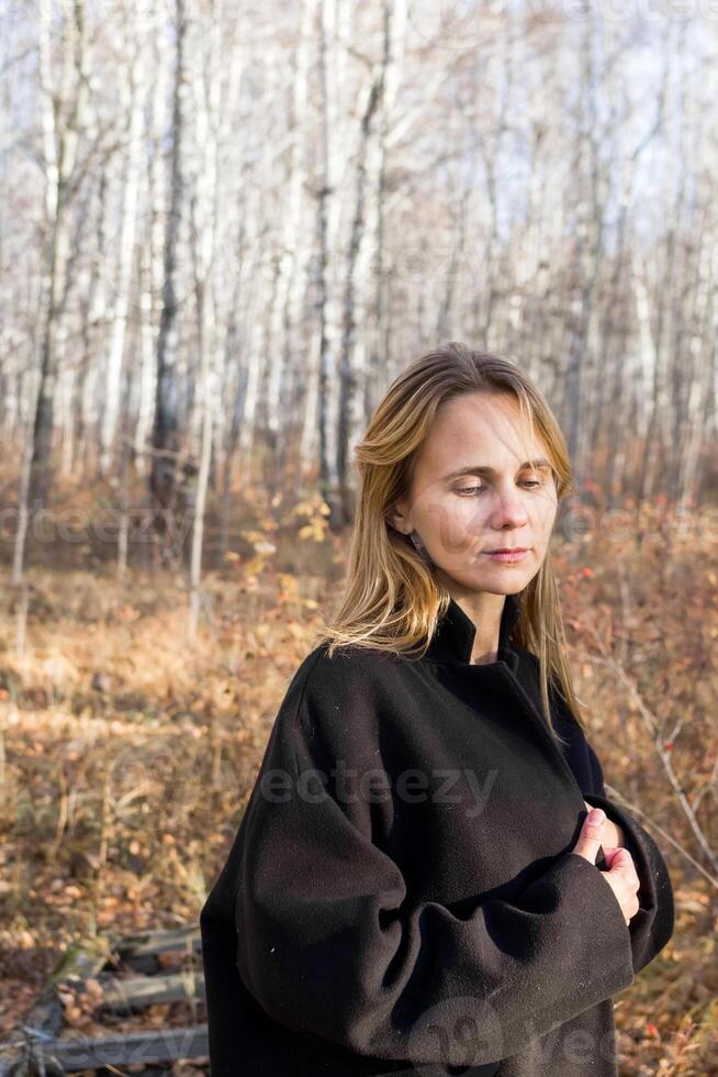 A young woman stands pensively in a dense forest. She is wearing a long black coat and has her hand on her chest. The sun is shining through the trees, creating a dappled pattern on the ground. photo