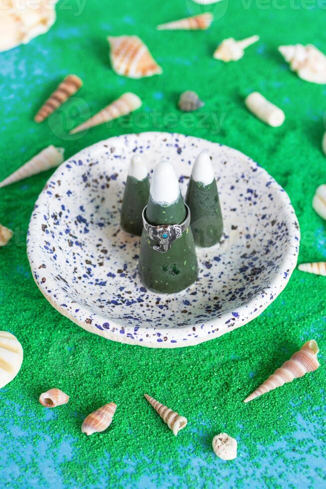 Handmade silver ring with blue stone on a ceramic ring holder and artificial green sand with seashells on a blue background. photo