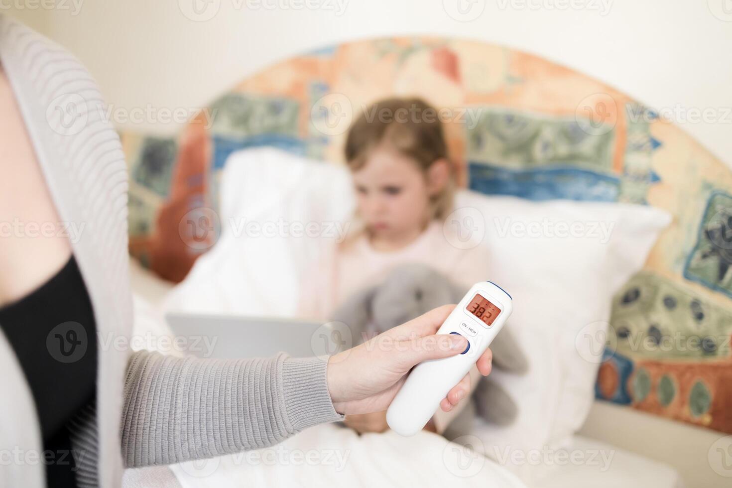 mother measuring temperature of her daughter. child got high temperature. Kid sitting in bed with tablet and furry toy and no face woman holding infrared thermometer which indicates 38,2 C photo