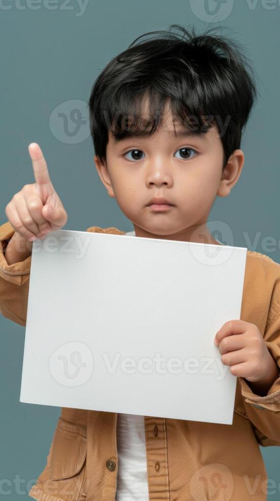 un pequeño tailandés chico alegremente sostiene arriba un blanco blanco papel, dando un pulgares arriba foto