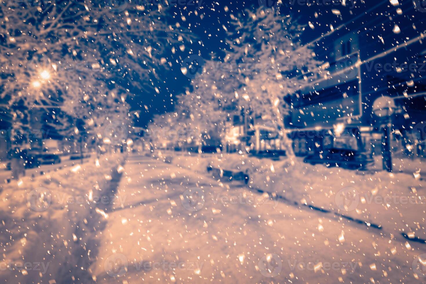 Blured photo of a winter night park with lanterns, pavement and trees covered with snow in snowfall.