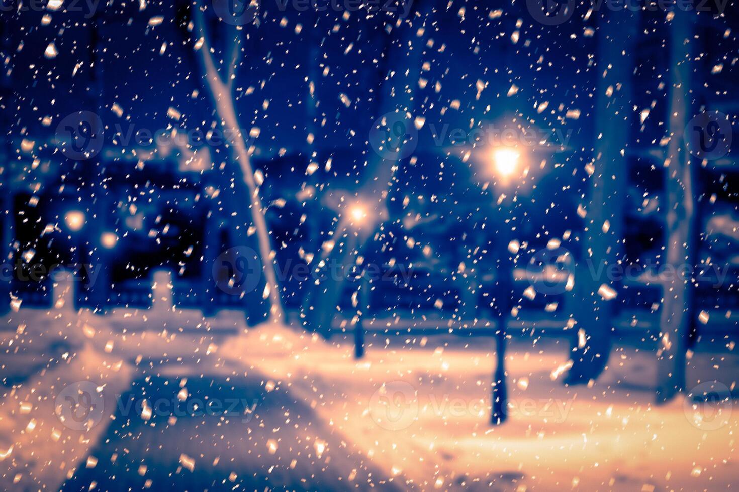 Blured photo of a winter night park with lanterns, pavement and trees covered with snow in snowfall.
