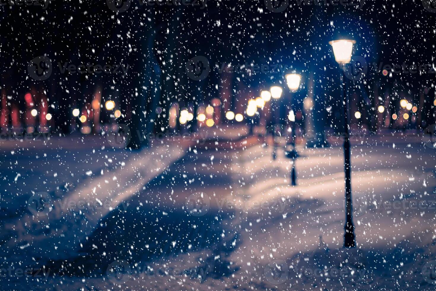 Blured photo of a winter night park with lanterns, pavement and trees covered with snow in snowfall.
