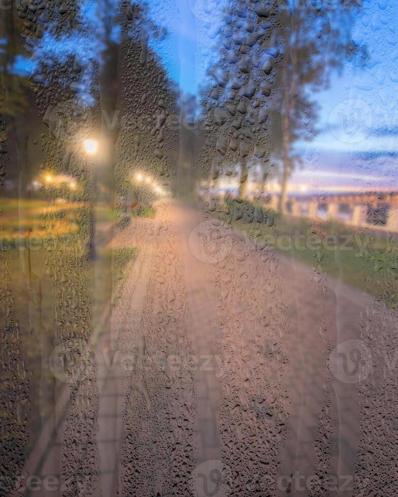 Defocused night park with lanterns behind rainy, wet glass at night. Concept of rainy autumn weather. photo