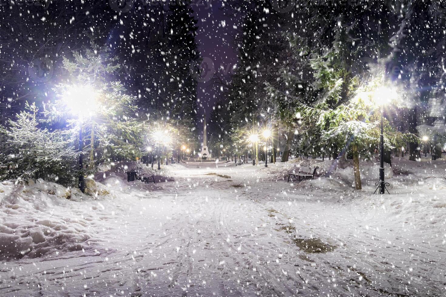Snowfall in a winter park at night with christmas decorations, lights and pavement covered with snow. photo