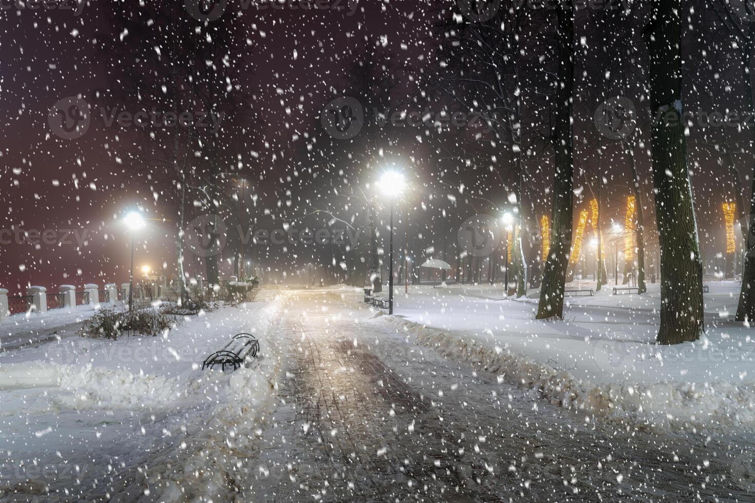 Snowfall in a winter park at night with christmas decorations, lights, pavement covered with snow and trees. photo
