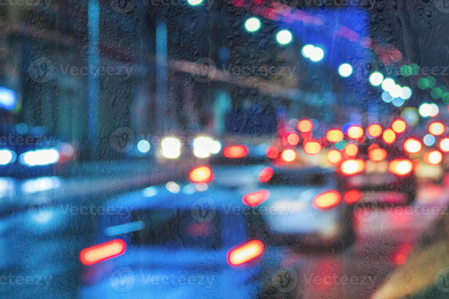 Blurry moving cars with headlights and lanterns reflecting on the wet asphalt in the night city behind rain-covered window. photo