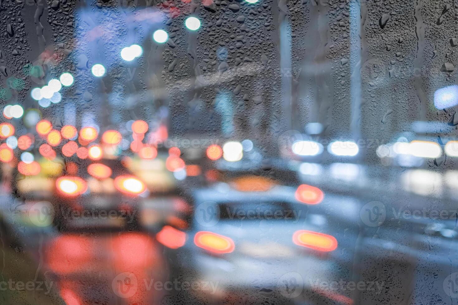 Blurry moving cars with headlights and lanterns reflecting on the wet asphalt in the night city behind rain-covered window. photo
