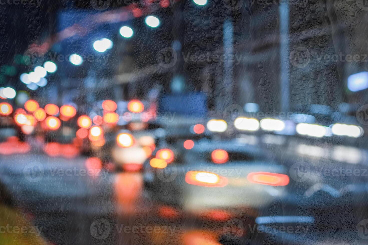 Blurry moving cars with headlights and lanterns reflecting on the wet asphalt in the night city behind rain-covered window. photo