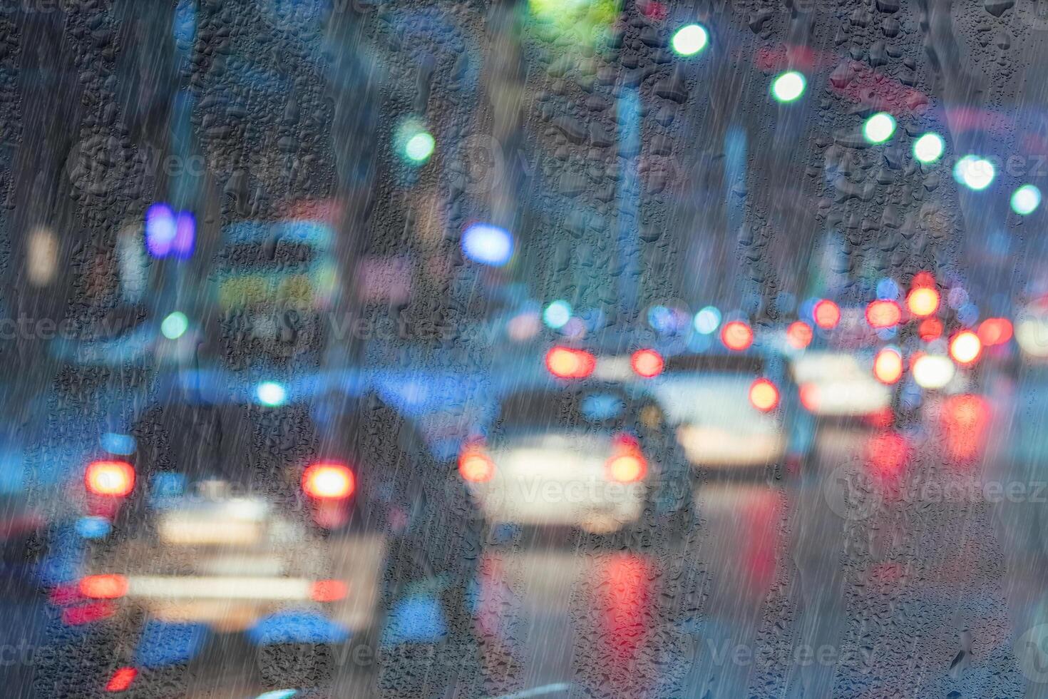Blurry moving cars with headlights and lanterns reflecting on the wet asphalt in the night city behind rain-covered window. photo