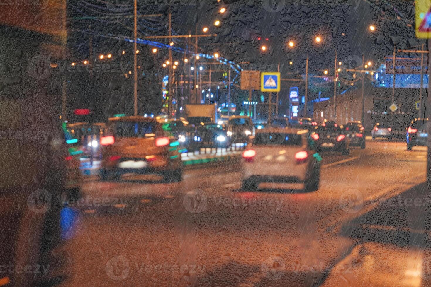 Blurry moving cars with headlights and lanterns reflecting on the wet asphalt in the night city behind rain-covered window. photo