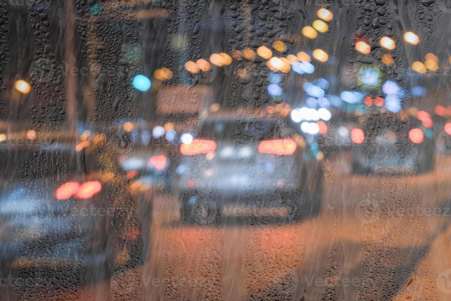 Blurry moving cars with headlights and lanterns reflecting on the wet asphalt in the night city behind rain-covered window. photo
